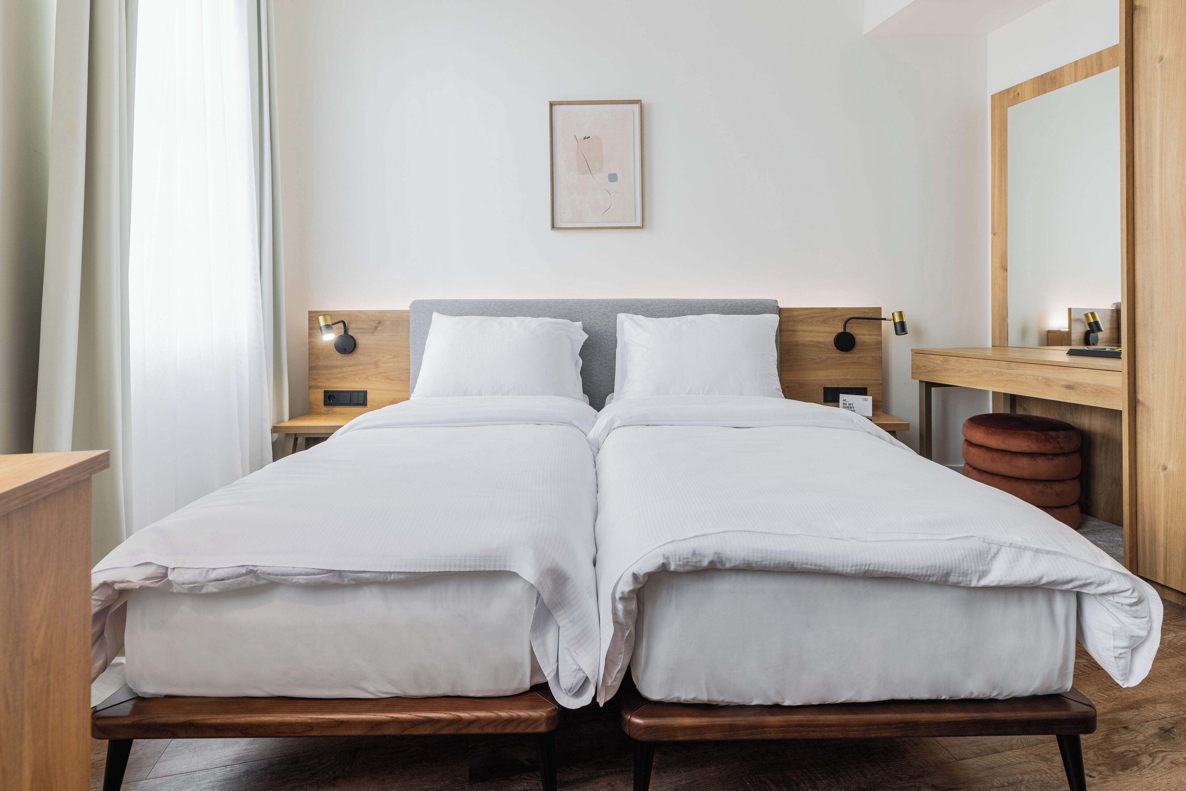 Uma cama de casal com roupa de cama branca, cabeceira de madeira, mesas de cabeceira com luminárias, um espelho e uma pequena obra de arte na parede.