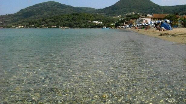 A beach with clear shallow waters, a sandy shore, and a backdrop of hills. - River Studios & Apartments (Corfu)