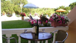 A patio with a round table, two wicker chairs, a bottle of Centenar wine, and two wine glasses. There are potted flowers and an umbrella in the background. - River Studios & Apartments (Corfu)