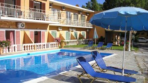 A pool area with lounge chairs and an umbrella, surrounded by a building with balconies and air conditioning units. - River Studios & Apartments (Corfu)