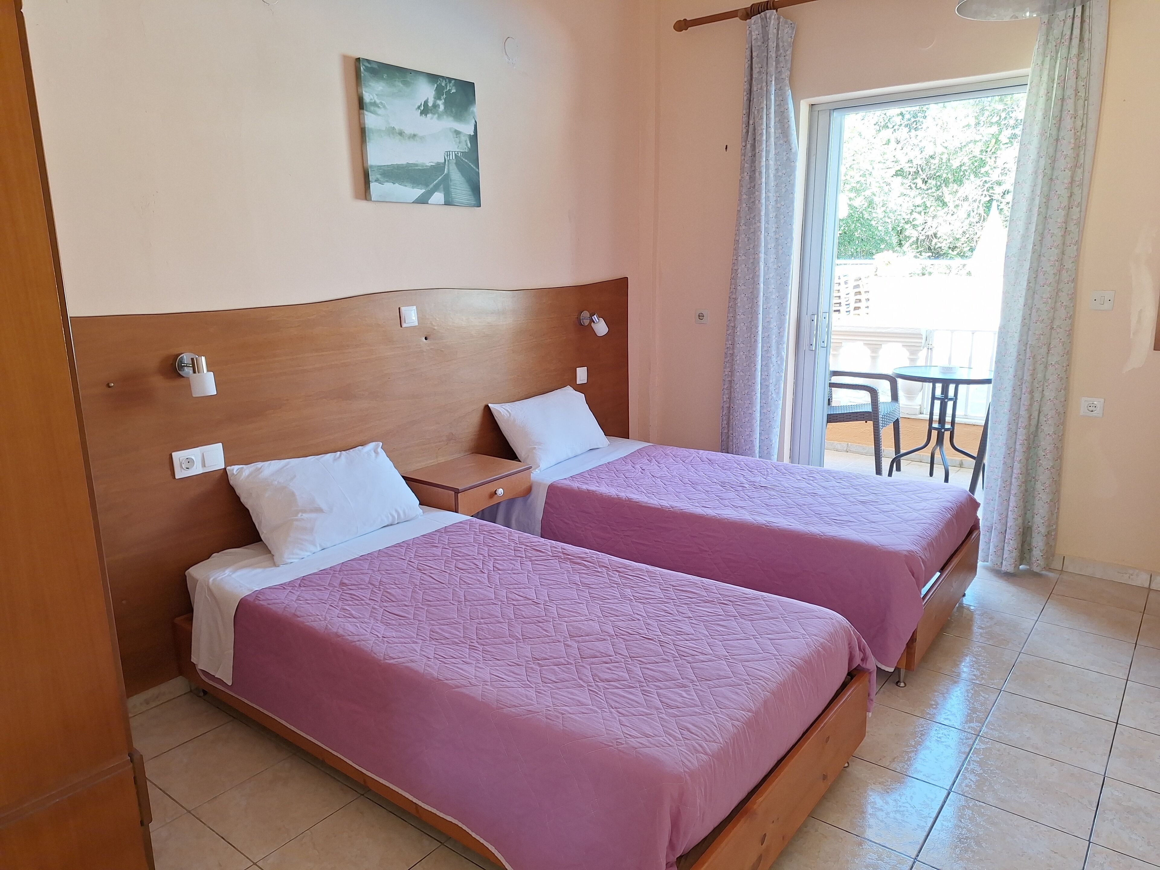 A hotel room with two beds, a wooden headboard, a painting on the wall, and a balcony with a table and chairs.