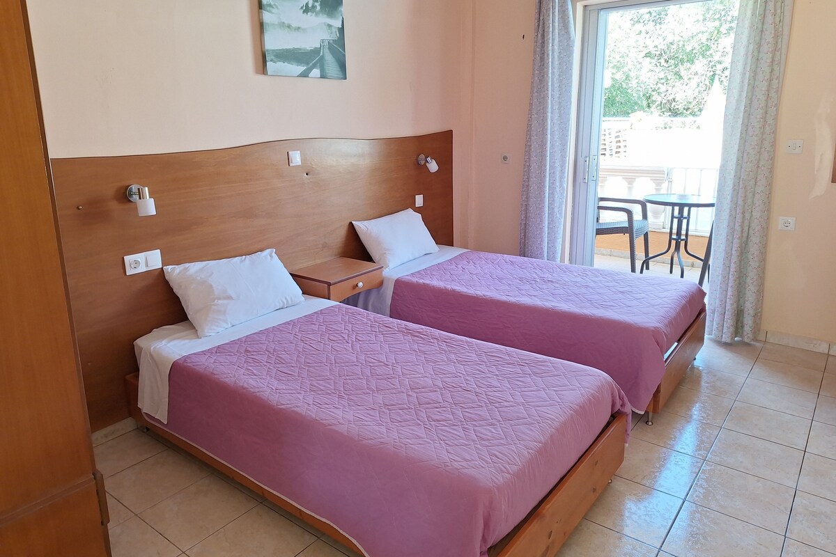 A hotel room with two beds, a wooden headboard, a painting on the wall, and a balcony with a table and chairs.