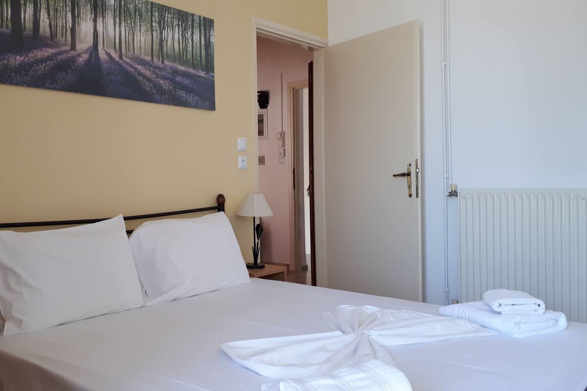 A neatly made bed with white linens and towels, a bedside lamp, and a painting of a forest scene on the wall.