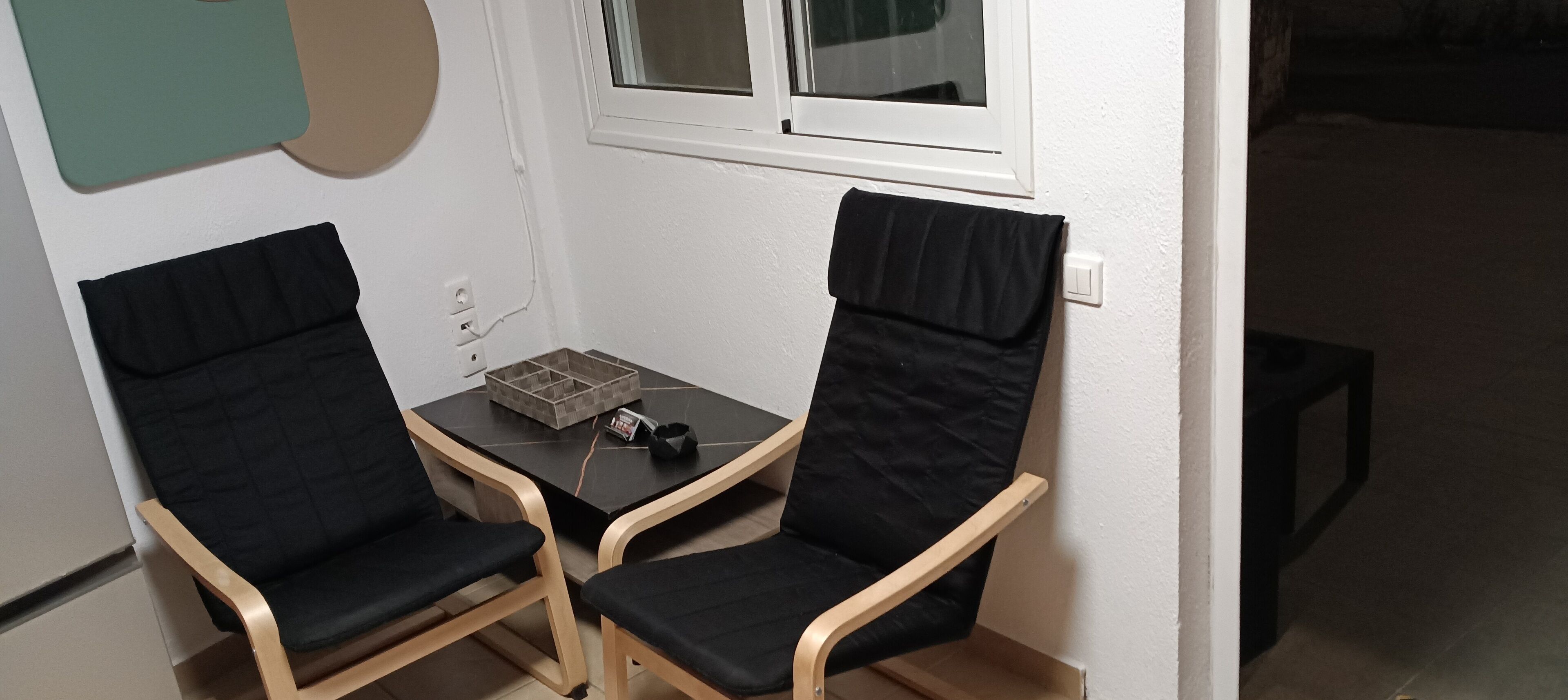 Deux fauteuils noirs à coussins, avec une structure en bois, une petite table sur laquelle se trouvent une boîte et une télécommande, et une fenêtre au-dessus.