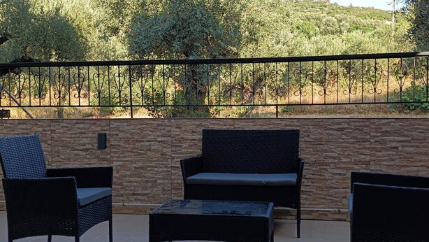 An outdoor patio with wicker furniture, a stone wall, and a view of a forested hillside. - Katerina Apartments (Kalamata)