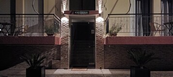 Entrada de hotel com uma placa indicando "KATERINA", fachada de pedra e vasos de plantas em ambos os lados.