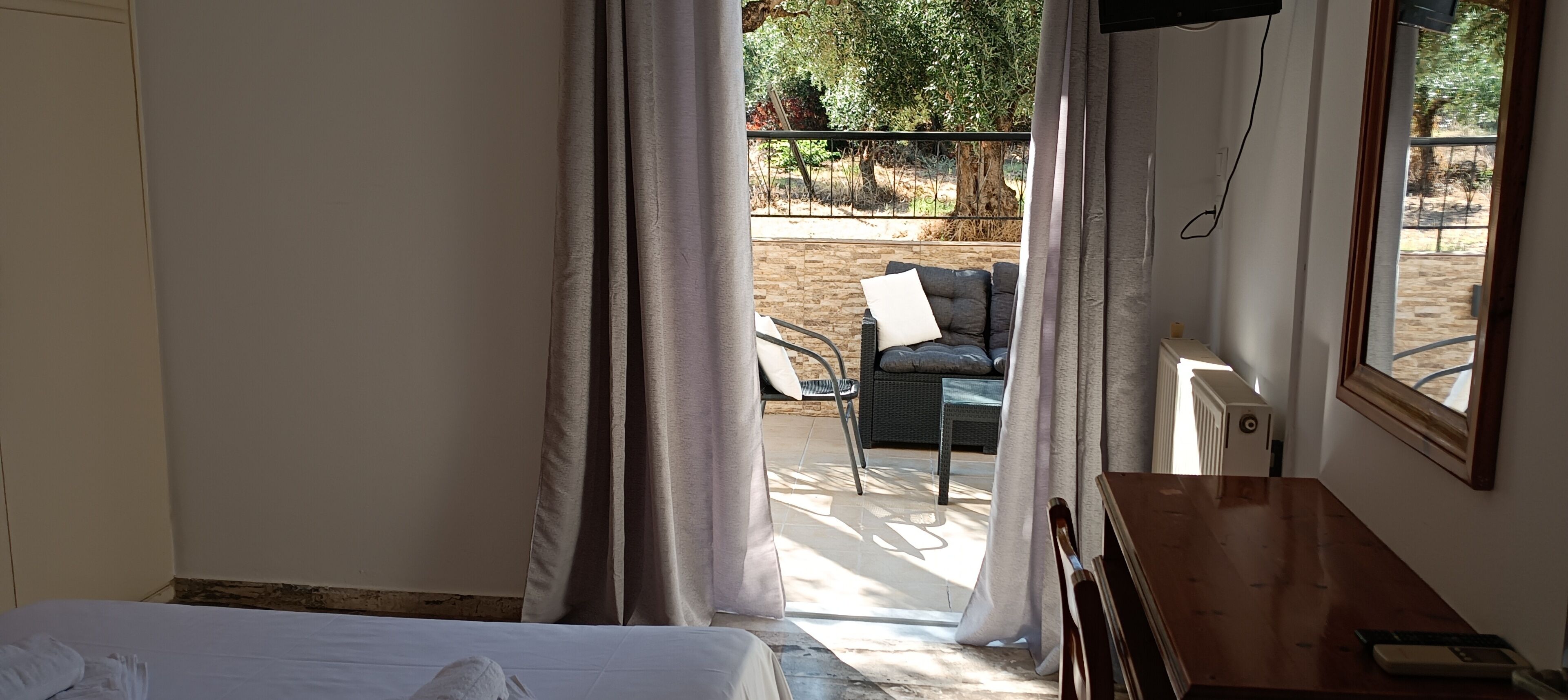 A hotel room with a bed, a wooden desk, a chair, and a view of an outdoor area with a patio and a tree.