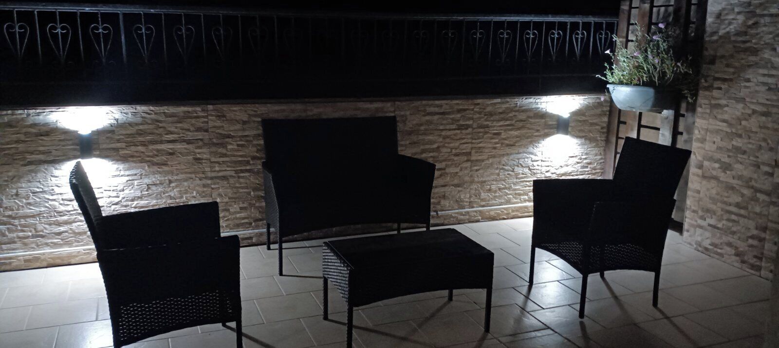 A patio area with three black wicker chairs and a small table, a hanging plant, and a stone wall backdrop.