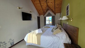 A hotel room with a wooden ceiling, a flat-screen TV, a bed with white bedding and a beige throw, and a window with curtains. - Cumbres Nevadas - Hostería Boutique (El Chalten)
