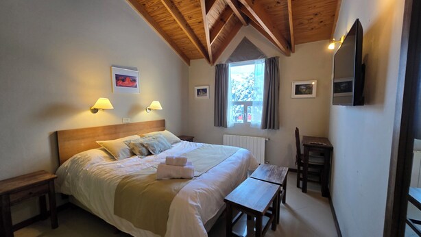 A bedroom with a wooden ceiling, a bed with white linens, two bedside lamps, a window with curtains, a small table, and a chair. - Cumbres Nevadas - Hostería Boutique (El Chalten)