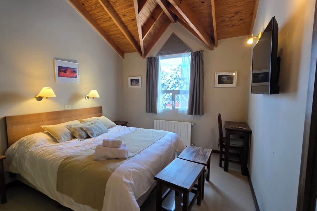 A bedroom with a wooden ceiling, a bed with white linens, two bedside lamps, a window with curtains, a small table, and a chair.