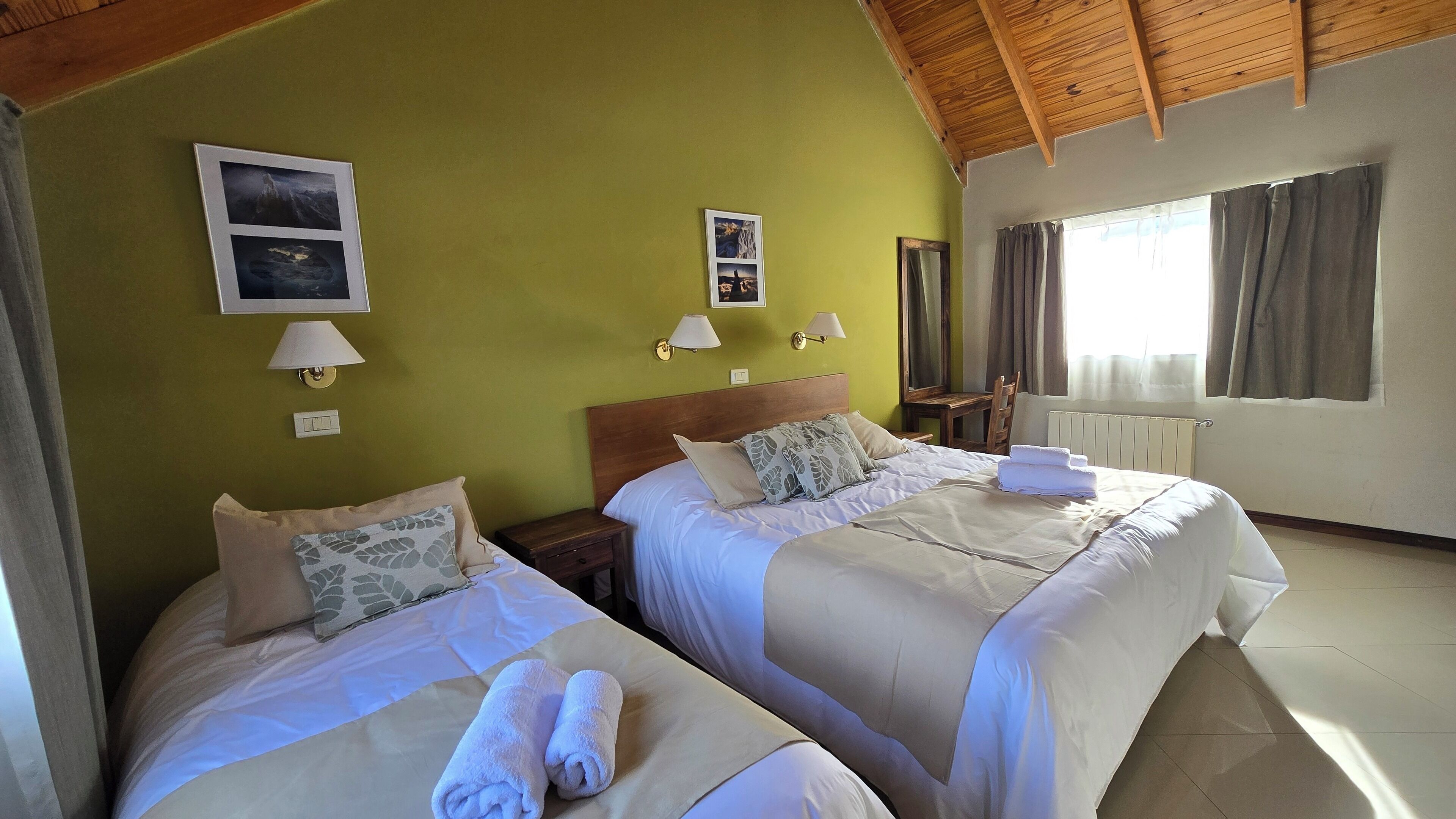 Habitación de hotel con dos camas, techo de madera, ventana con cortinas y lámparas fijadas a la pared.