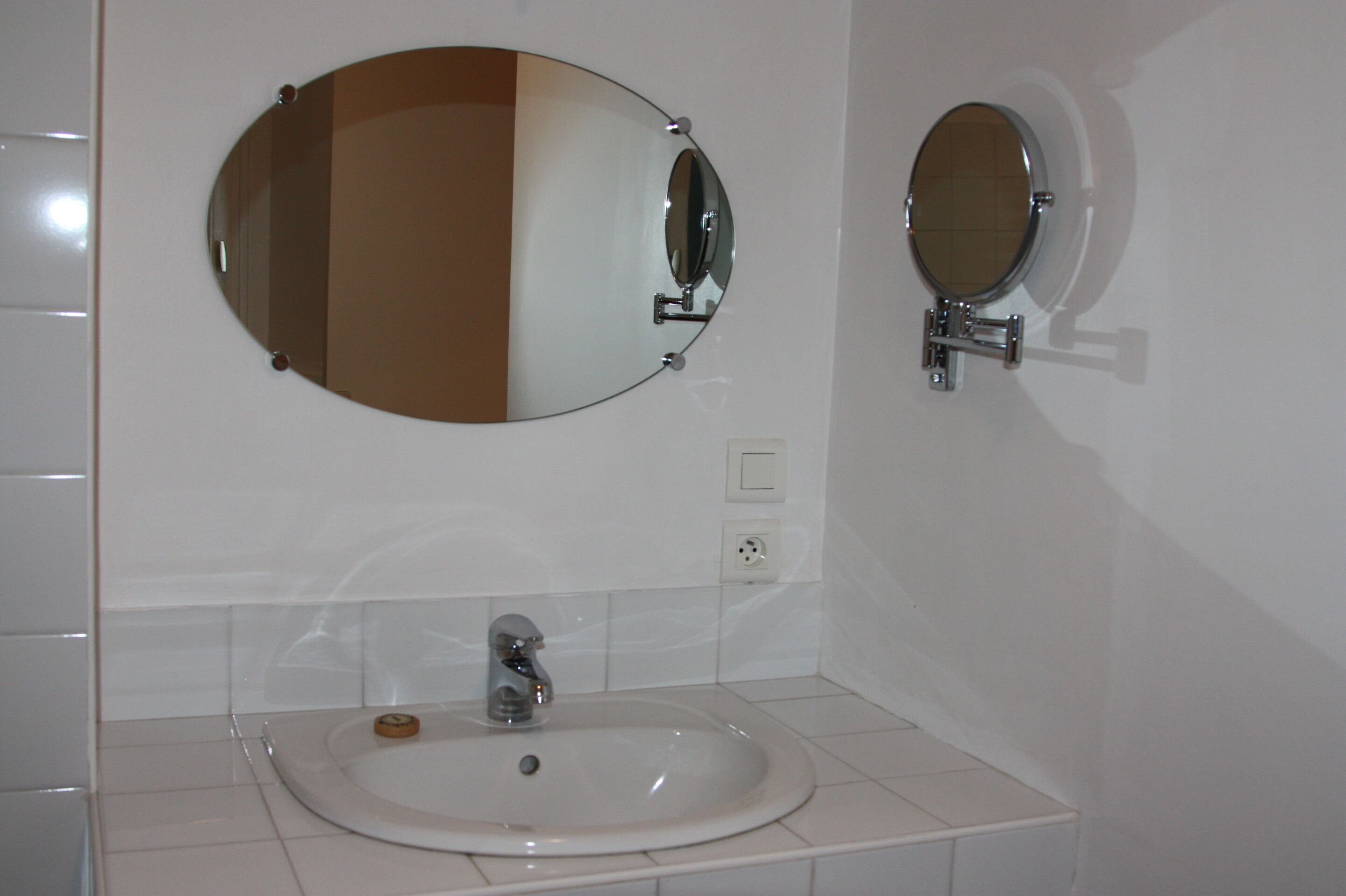 Un baño con un lavamanos blanco, un espejo ovalado grande y un espejo redondo más pequeño.