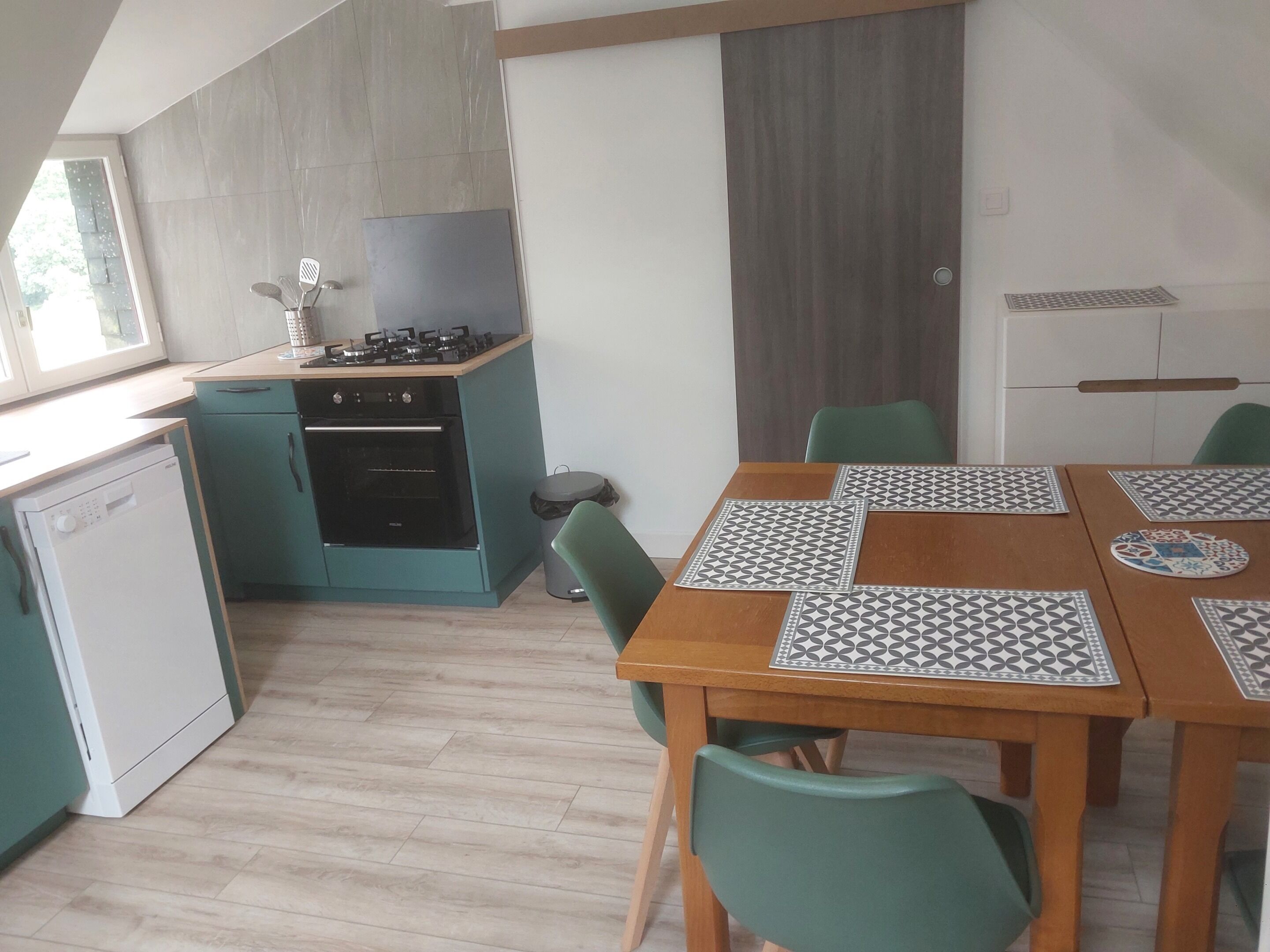 A compact kitchen with a gas stove, oven, and dishwasher. There is a dining table with patterned placemats and green chairs.