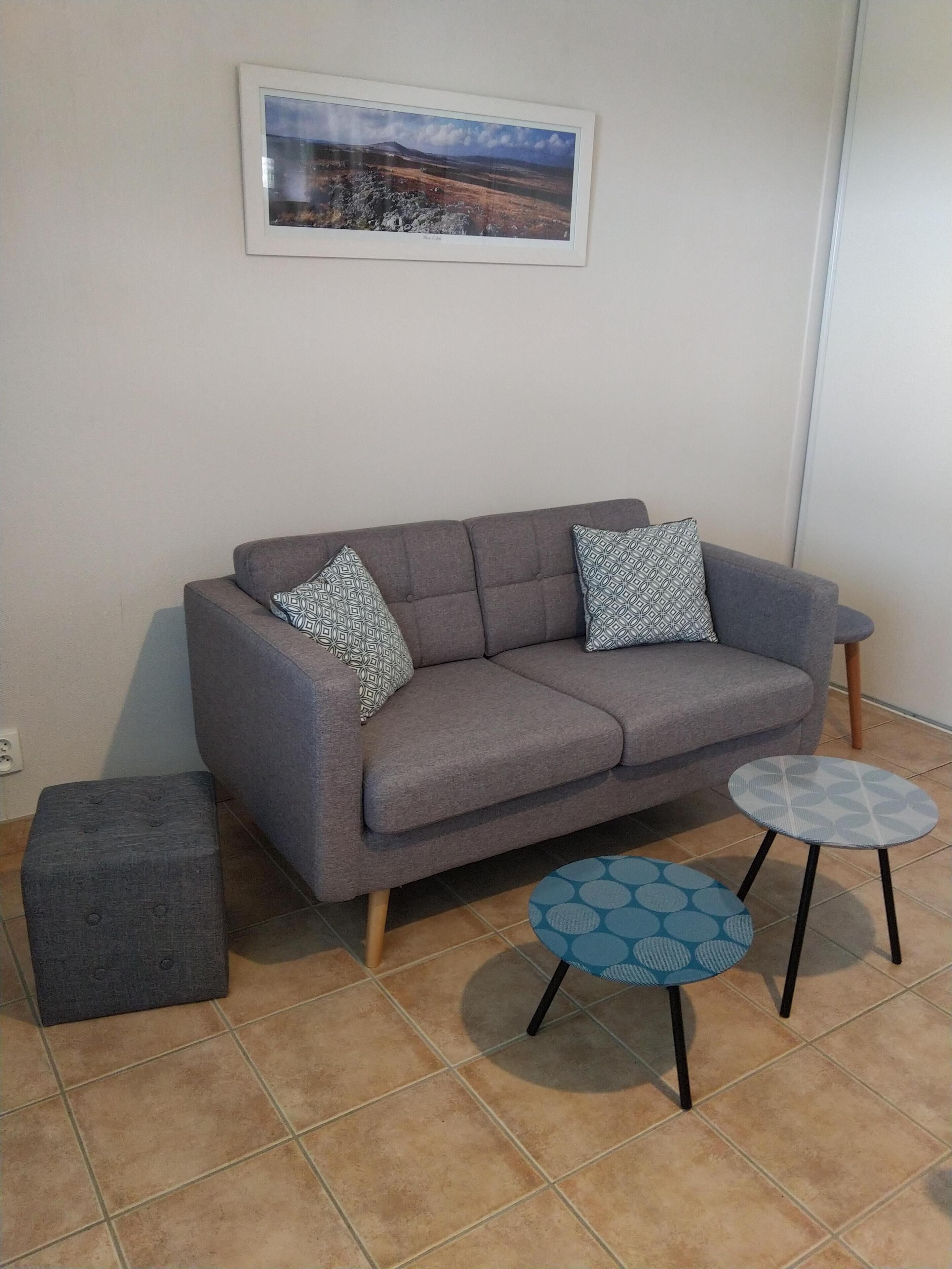 Ein graues Sofa mit gemusterten Kissen, ein grauer Hocker und ein kleiner runder Tisch in Blau und Weiß mit schwarzen Beinen.