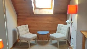 Un dormitorio acogedor en el ático, con techo inclinado, tragaluces, dos sillones tapizados, una pequeña mesa redonda y una lámpara de noche.