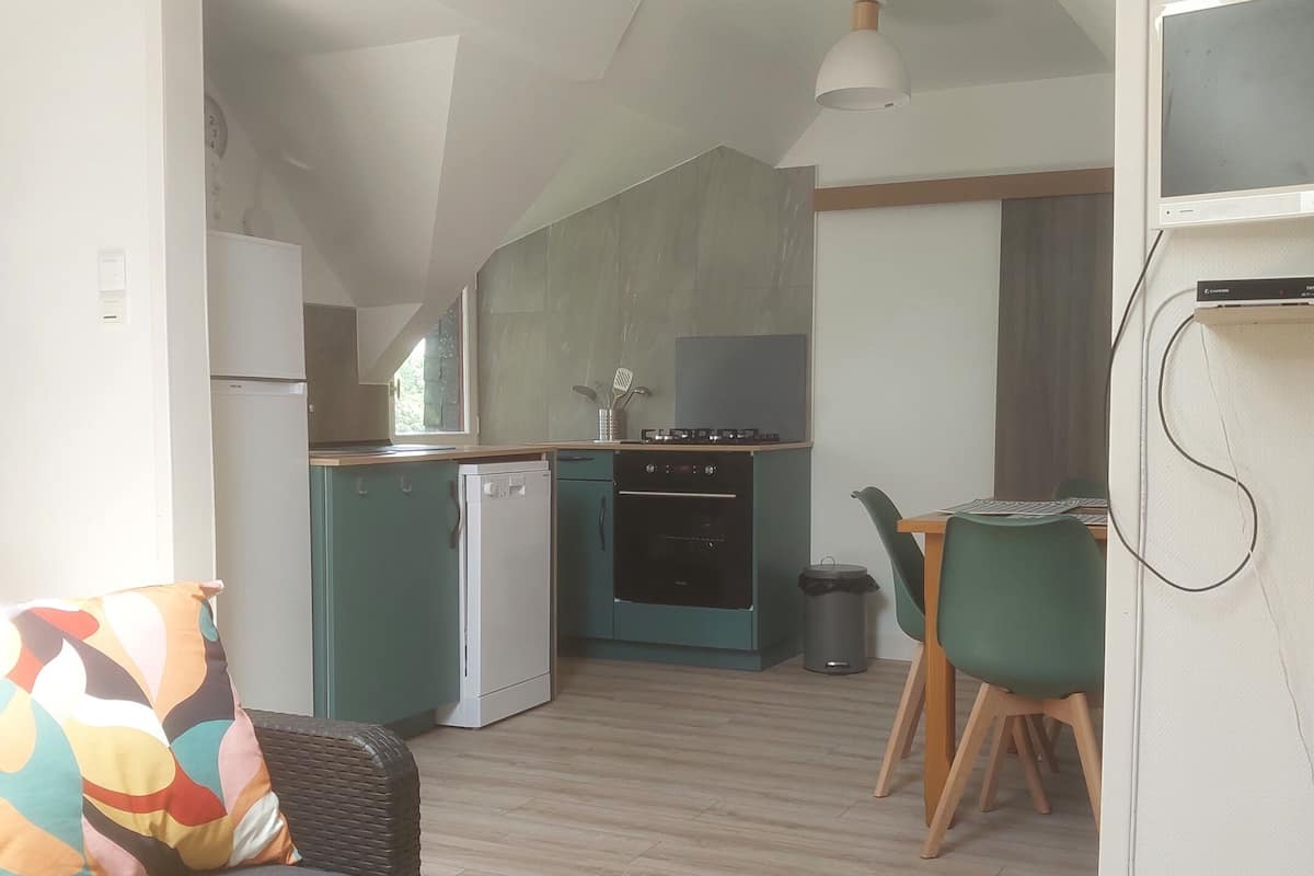 A compact kitchen with a refrigerator, stove, and sink. There is a dining area with a table and chairs, and a sofa with a colorful cushion.