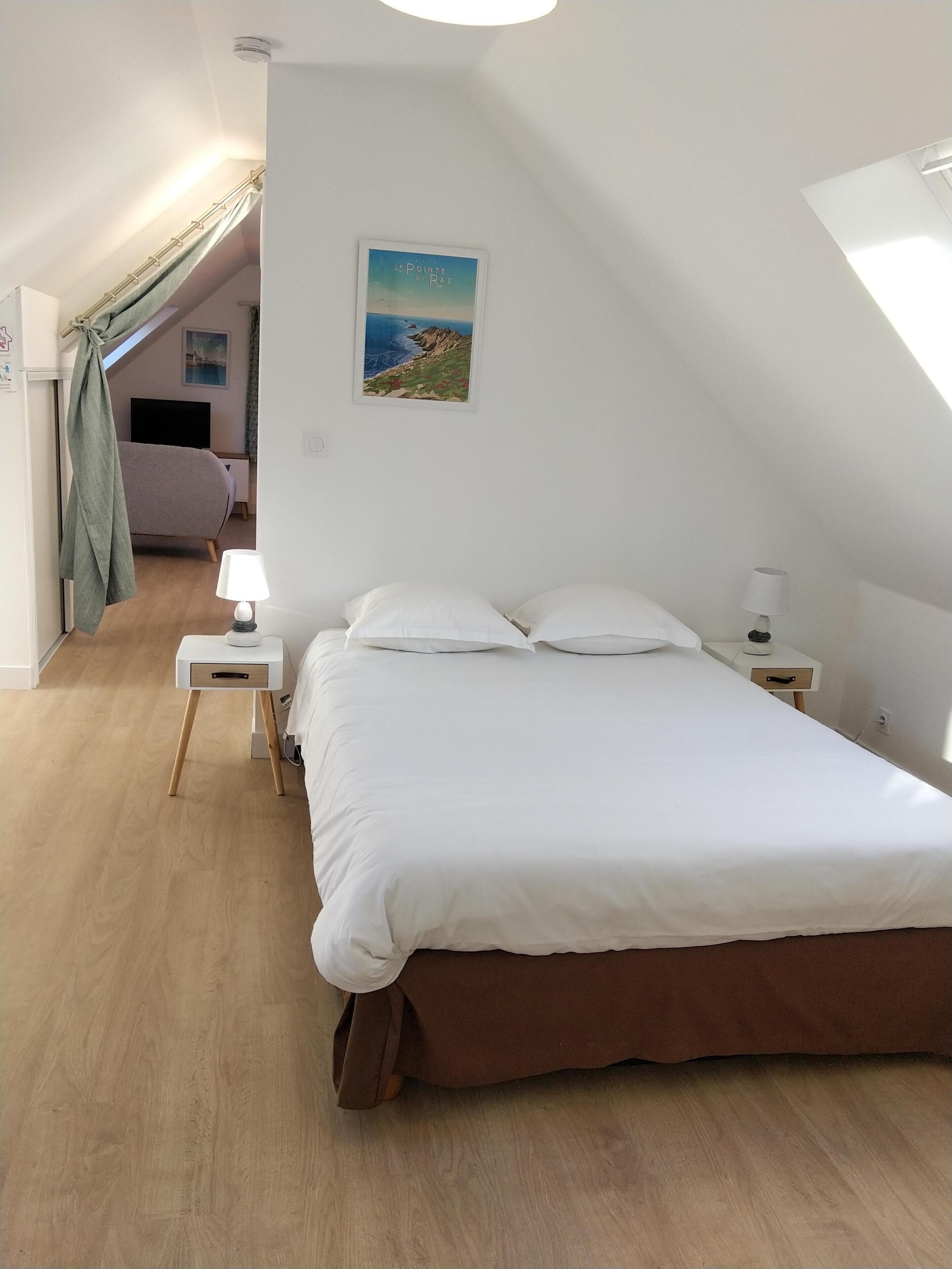 A bedroom with a large bed, a bedside table with a lamp, a small drawer, and a wall-mounted picture.