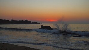 A sunset over a coastal area with waves crashing onto the shore and a silhouette of buildings in the background. - Hotel Arboleda (Sanxenxo)