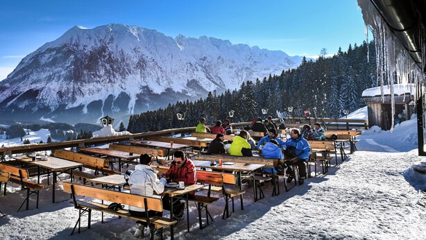 Un espacio al aire libre cubierto de nieve con bancos y mesas de madera, con vistas a una cadena montañosa y un valle boscoso.