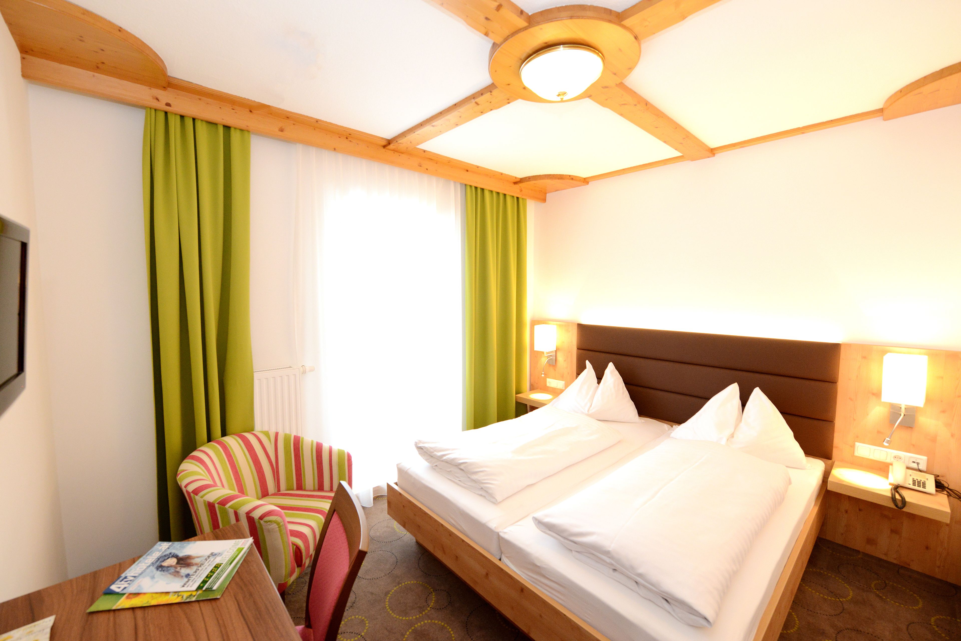 A hotel room with a wooden ceiling, a bed with white linens, a striped armchair, a small table with a magazine, and a window with green curtains.