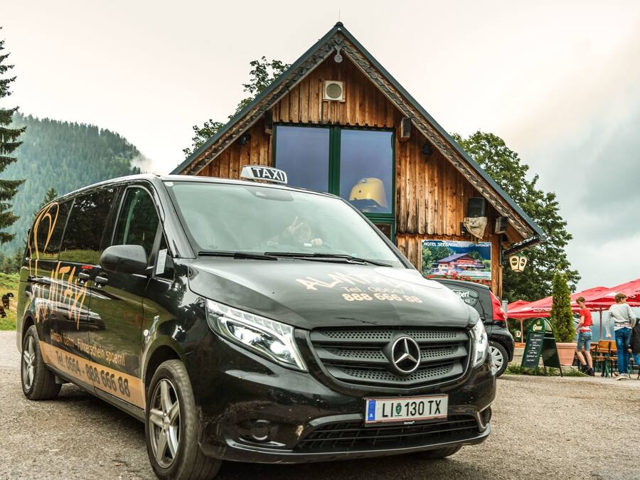 A black van with a 'TAXI' sign parked in front of a wooden building with a sign that reads 'HOTEL'.