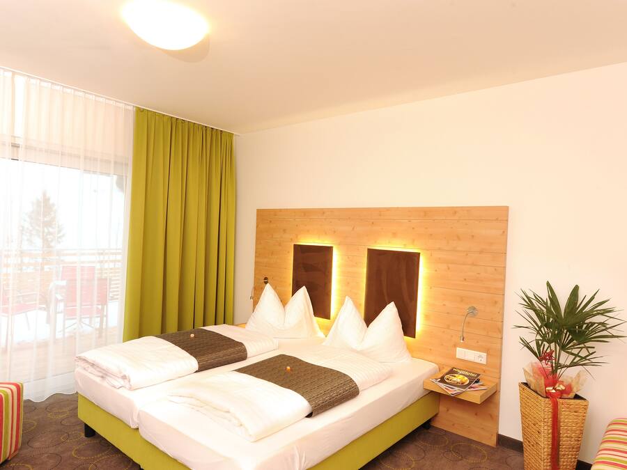 A hotel room with a large bed, a wooden headboard with two wall-mounted lights, a green curtain, a small bedside table with a magazine, and a potted plant.