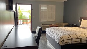 A hotel room with a bed, a desk, a chair, a window with blinds, and a view of greenery outside. - La Casa Inn (Pasadena)
