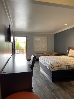 A hotel room with a bed, a desk, a chair, a window with blinds, and a view of greenery outside.