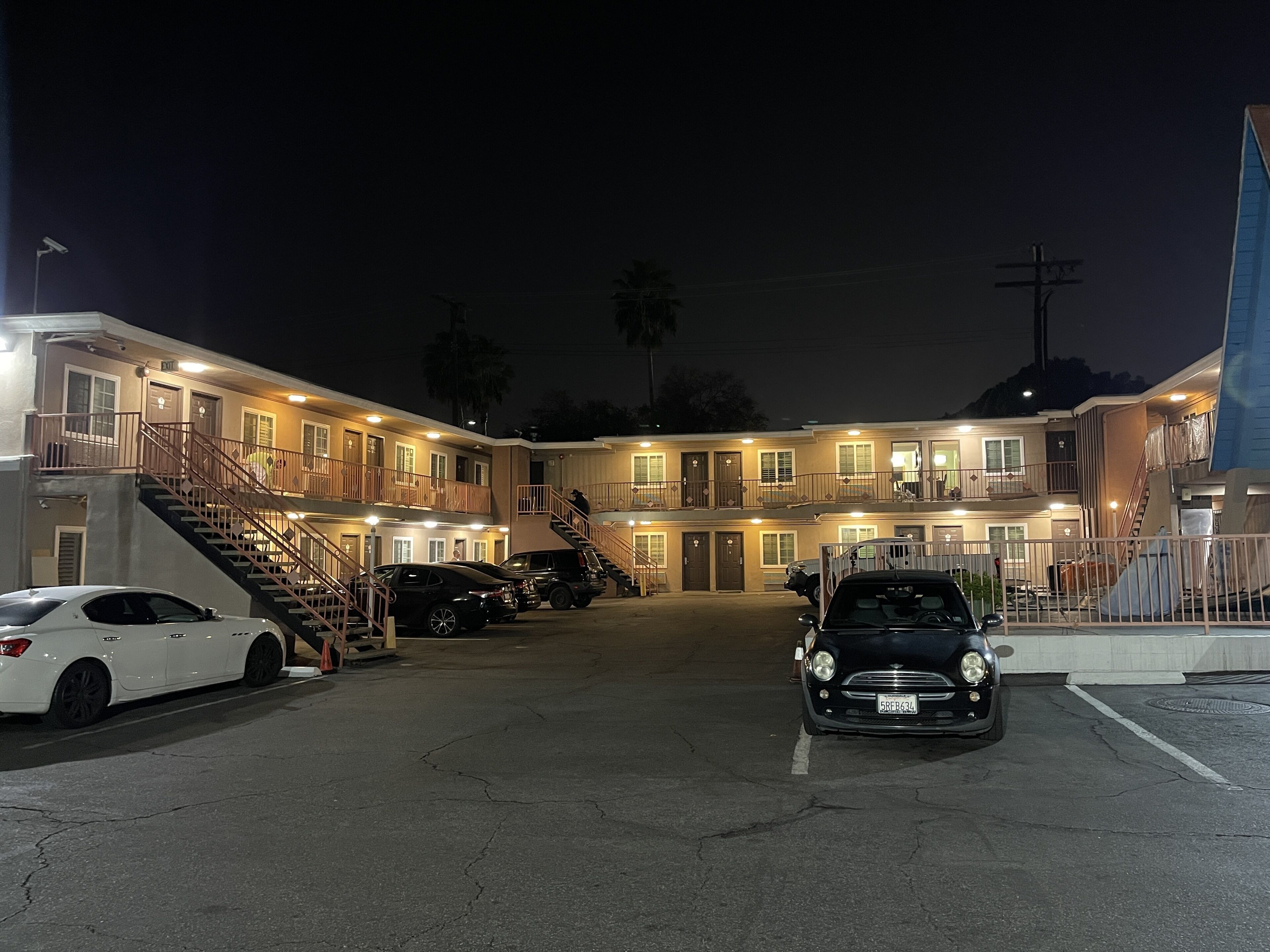A multi-story motel with a central entrance, a parking area with several cars, and a staircase leading to the upper floors.