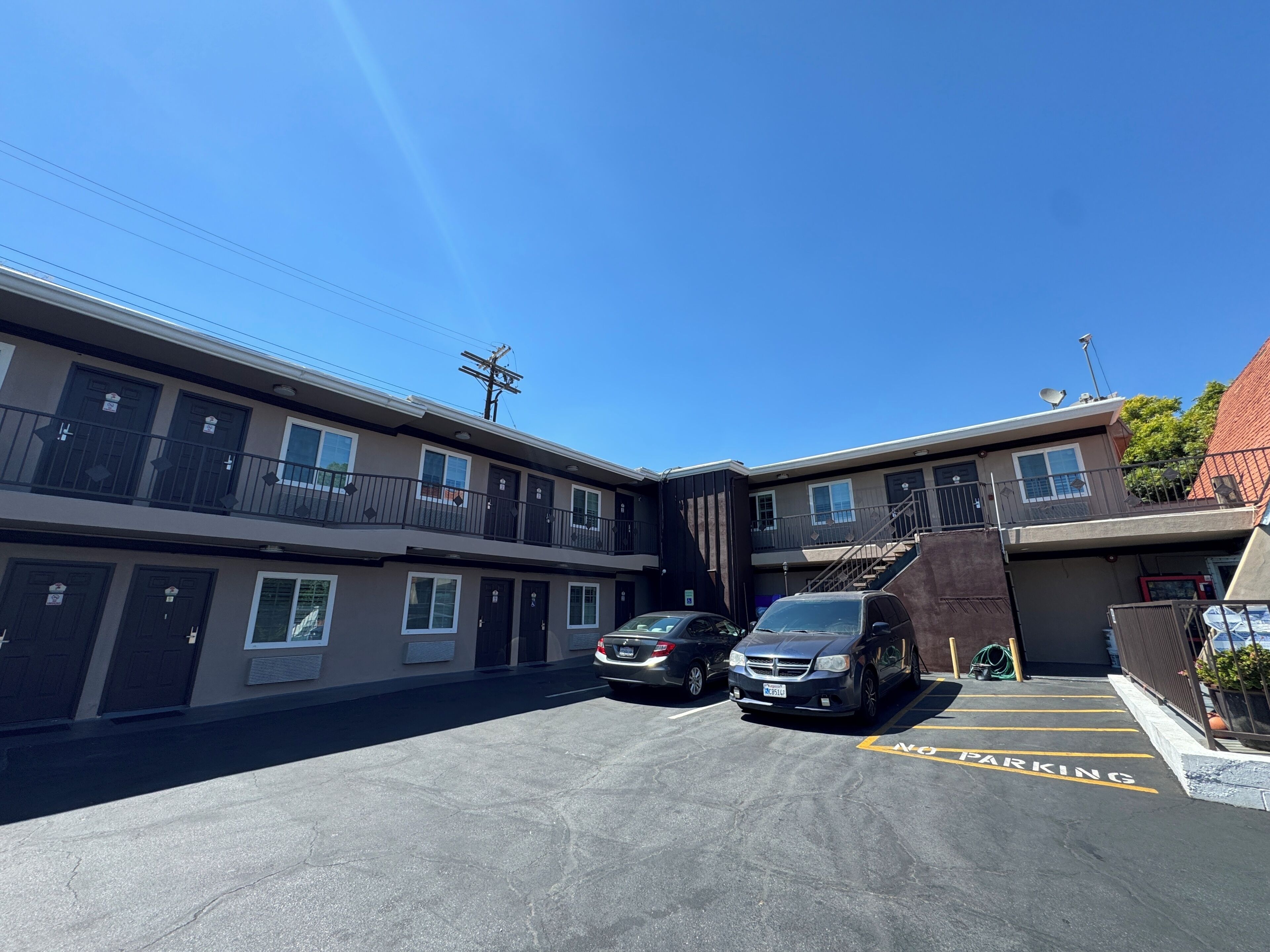 A multi-story motel with parking spaces, a clear blue sky, and a red-roofed building in the background.