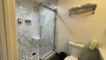 Un baño con ducha envuelta en vidrio, inodoro, lavamanos con toallero y un cesto de basura.