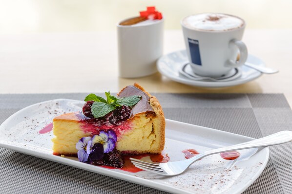 A slice of cheesecake with berries and mint on a white plate, a fork, and a cup of coffee in the background.