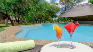 A poolside setting with two drinks on a table, a thatched-roof structure, and lush greenery in the background. - Mongena Game Lodge (Hammanskraal)