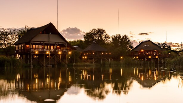 Three wooden structures with thatched roofs, elevated on stilts over a calm body of water, reflecting the structures and lights. - Mongena Game Lodge (Hammanskraal)