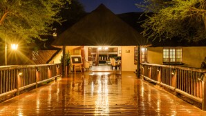 A wooden deck leading to a building with a thatched roof, surrounded by trees and illuminated by outdoor lights. - Mongena Game Lodge (Hammanskraal)