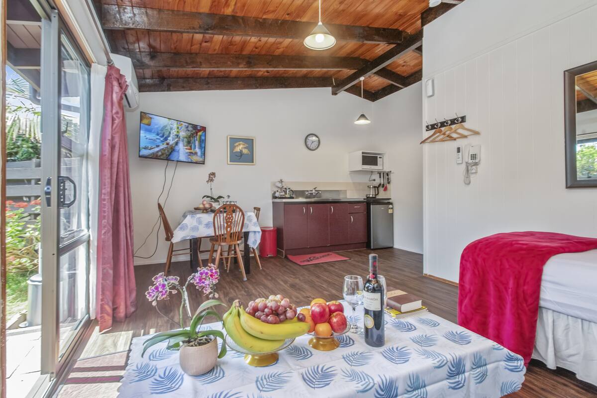 A compact room with a bed, a dining table with fruit, a kitchen area, and a TV.