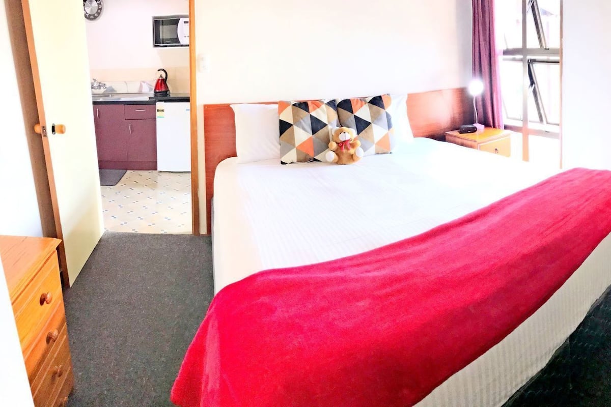 A hotel room with a bed, a nightstand, a lamp, a window, and a view of the kitchen.