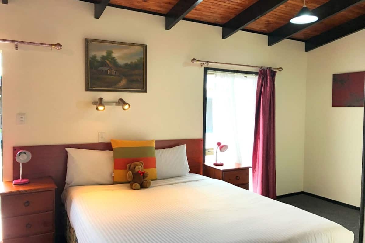 A bedroom with a bed, a nightstand, a lamp, a painting, and a window with curtains.