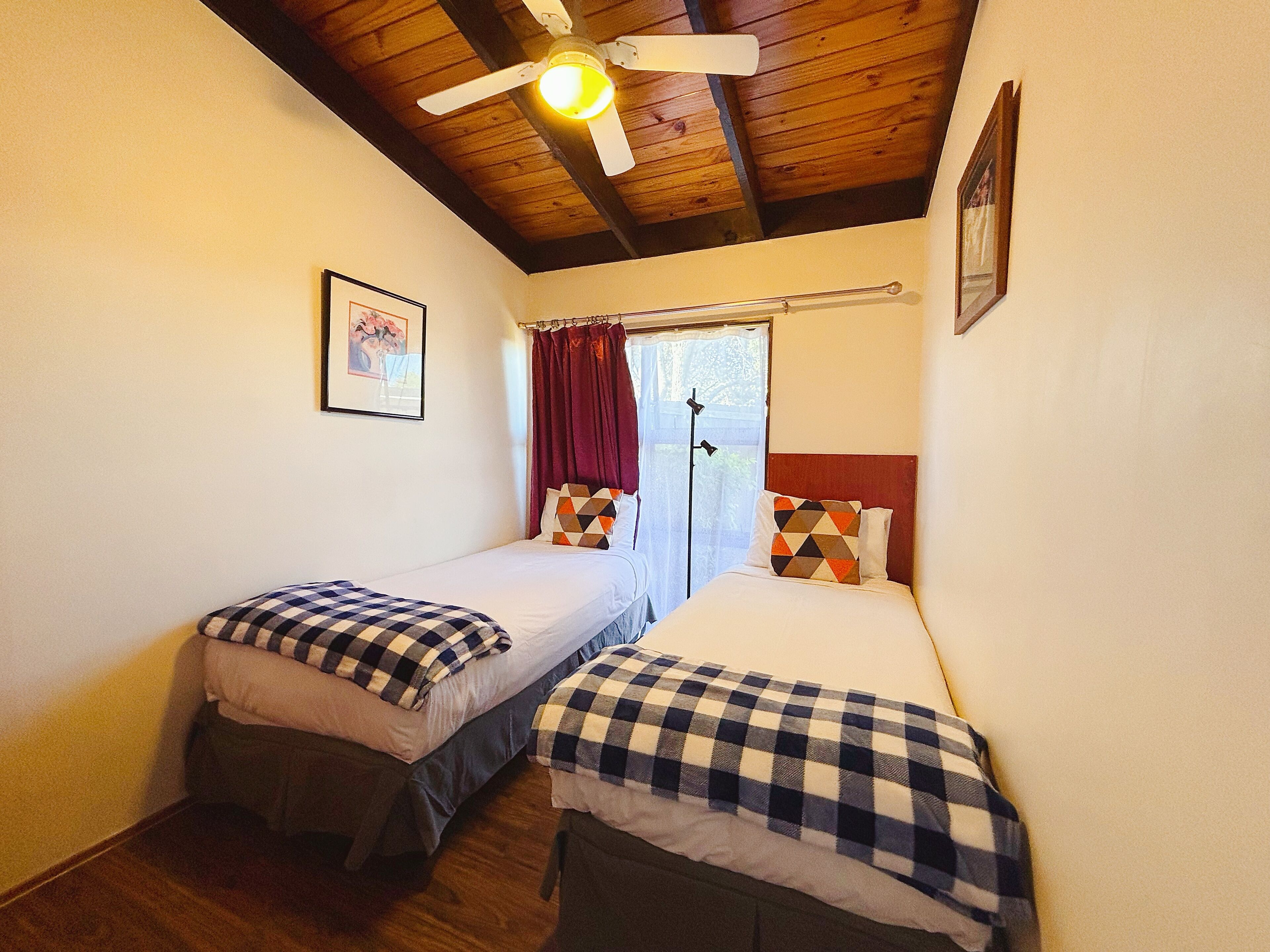 A room with two single beds, a ceiling fan, a window with curtains, and a framed picture on the wall.