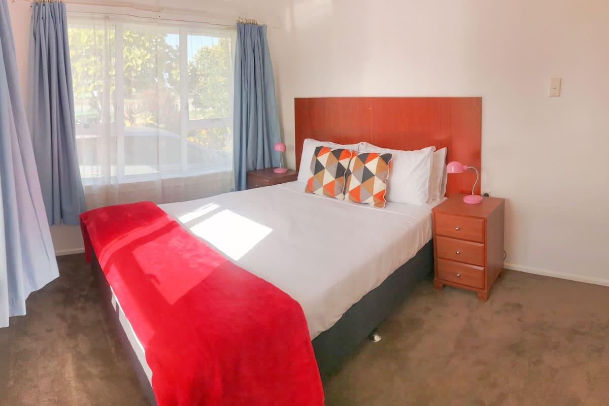 A neatly made bed with a red blanket, a wooden nightstand with a lamp, and a window with curtains.
