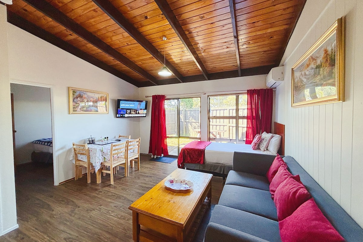 A cozy room with a wooden ceiling, a bed, a sofa, a dining table with chairs, and a TV mounted on the wall.