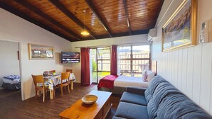 A cozy room with a wooden ceiling, a sofa, a dining table with chairs, a TV, and a window with curtains. - Grange Motel (Auckland)
