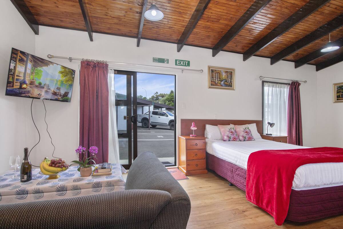 A hotel room with a bed, a sofa, a table with a bottle and fruit, a TV, and an exit sign.