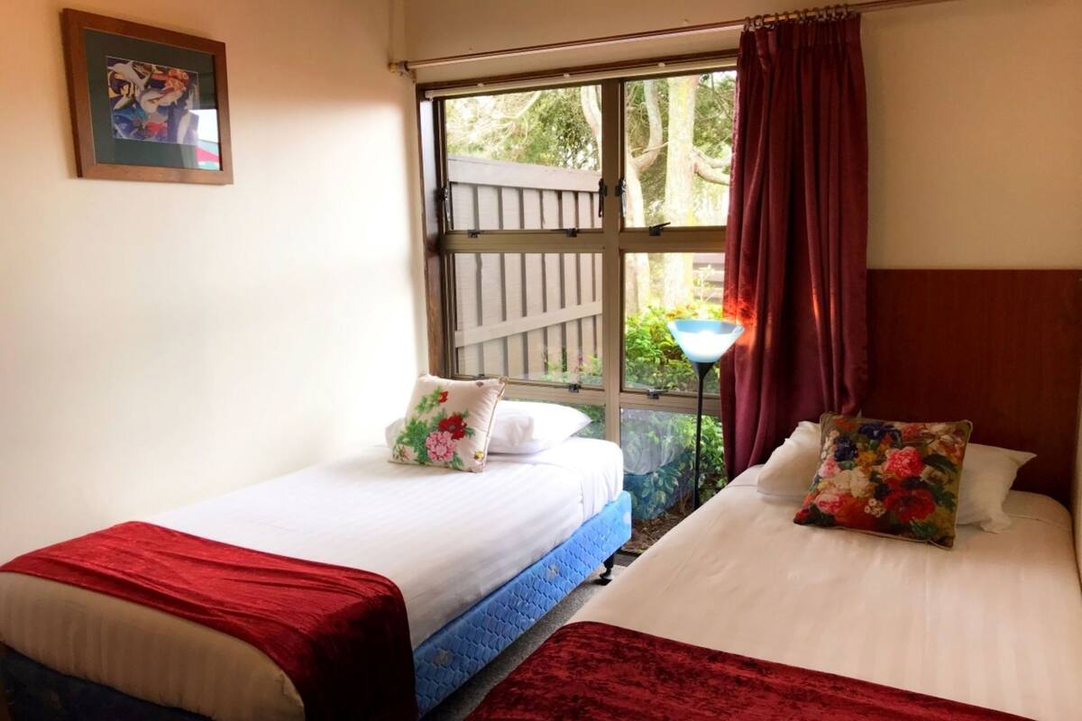 A room with two beds, a window with curtains, a framed picture on the wall, and a nightstand with a lamp.