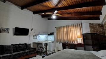 Ein Zimmer mit Holzboden, Deckenventilator, einem Bett, einem Schlafsofa, einem Schreibtisch mit Stuhl, einem an der Wand montierten Fernseher und einem Fenster mit Vorhängen.