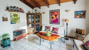 A living room with a wooden ceiling, tiled floor, a sofa with colorful cushions, a wooden coffee table, a cabinet with glass doors, a lamp, and a radio on a side table. - Dai Nonni Hotel (Guatemala City)
