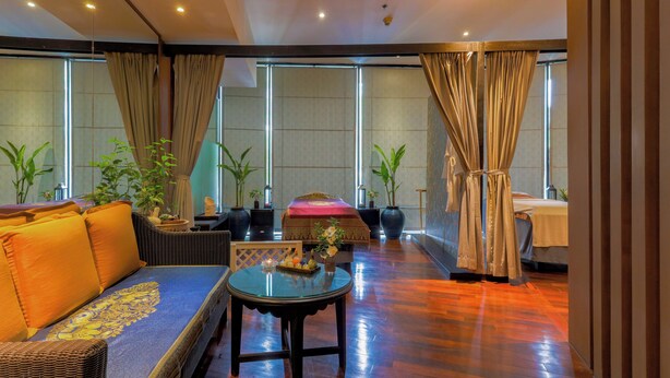 A spacious room with a large sofa, a round table, and a bed. There are potted plants and curtains.