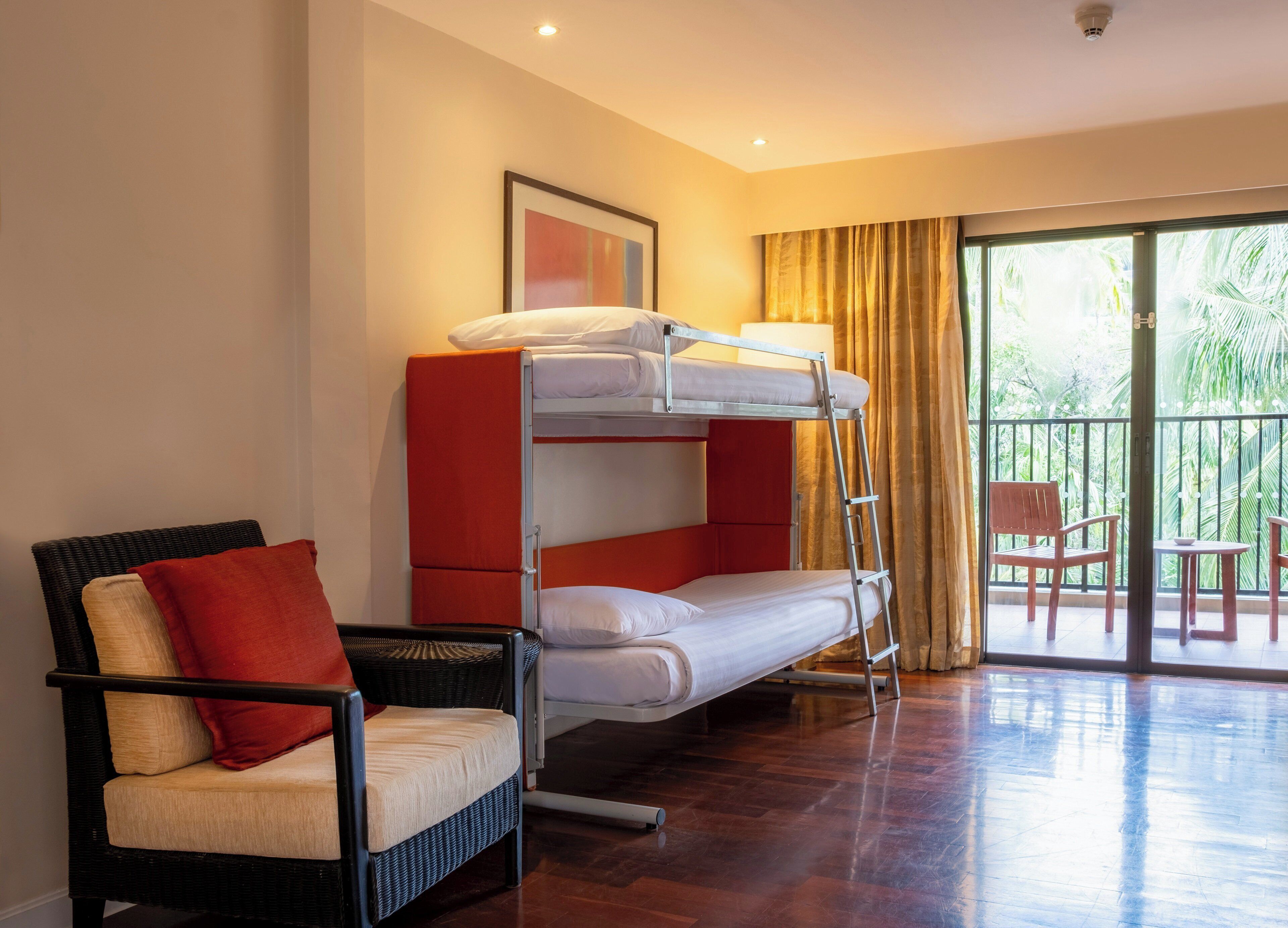 Une chambre d’hôtel avec un lit superposé, une chaise en osier avec un coussin, une petite table et une vue sur les palmiers par la fenêtre.