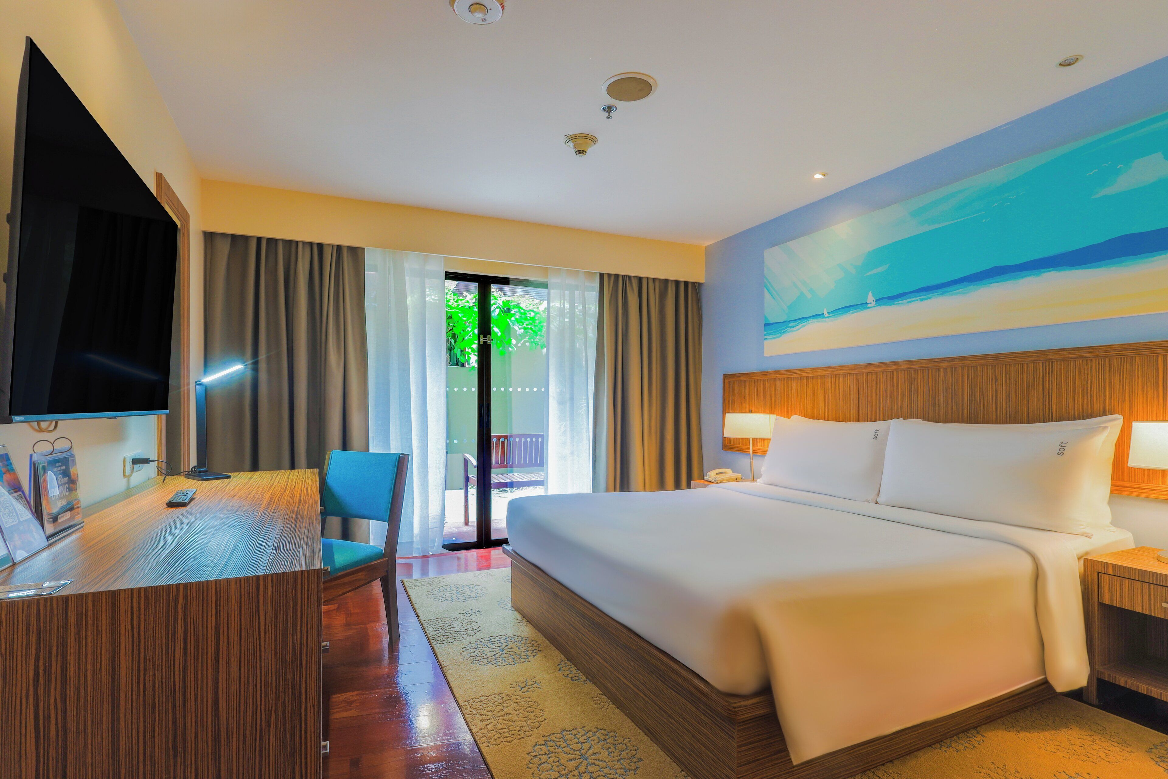 A hotel room with a large bed, a desk with a computer, a TV, and a view of the outdoors through a glass door.